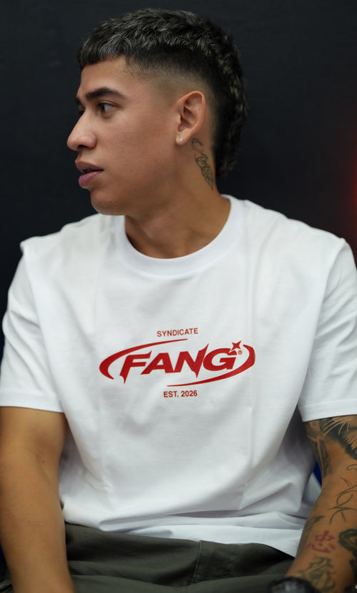 barbero sentado con una camiseta fang sello en blanco y rojo. lleva tatuajes en el cuello y el brazo. esta observando el trabajo de su compañero. 