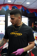 barbero trabajando con una camiseta fang sello en negro y dorado. lleva cadenas de oro. la barbería es colorida en rojo y azul.