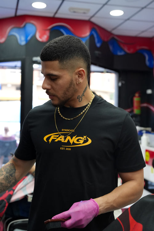 barbero trabajando con una camiseta fang sello en negro y dorado. lleva cadenas de oro. la barbería es colorida en rojo y azul.